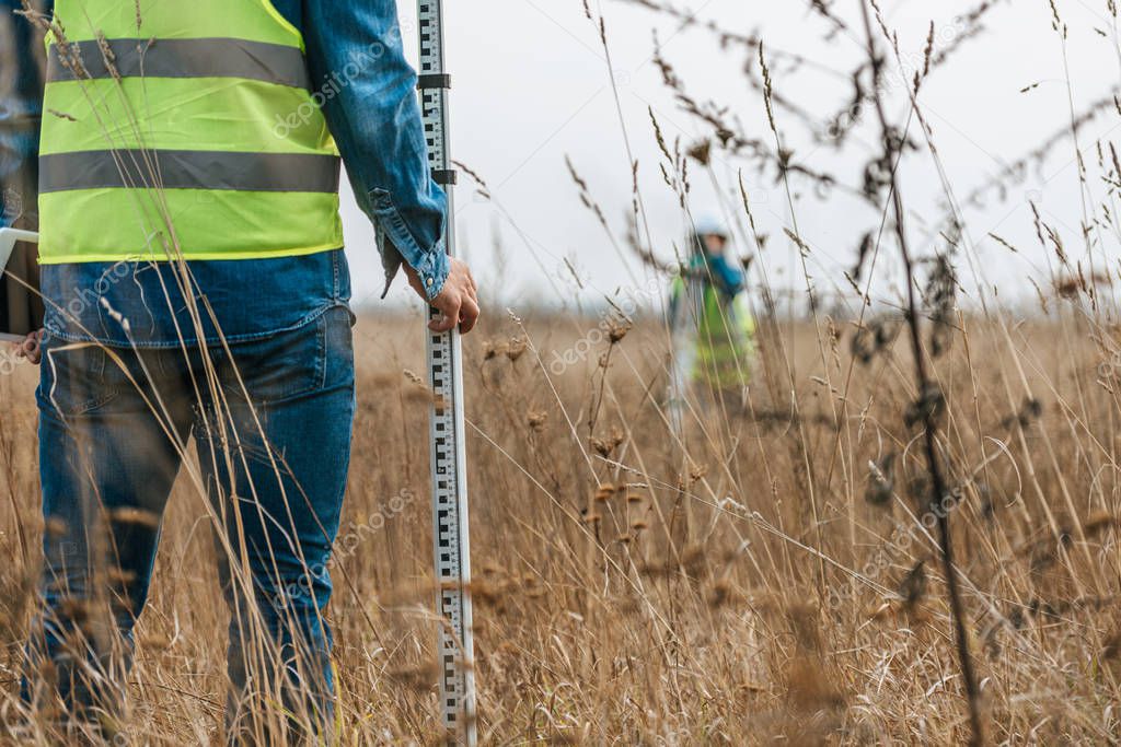 Selective focus of surveyor with ruler and colleague with digital level in field