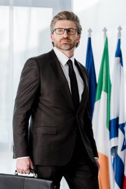 Yakışıklı sakallı diplomat bayrakların yanında evrak çantası tutuyor. 