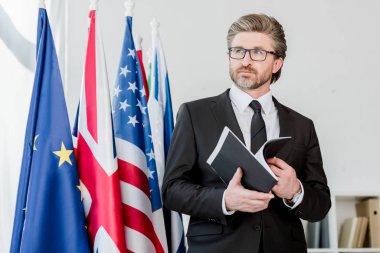 Bayrakların yanında klasör tutan sakallı diplomat. 