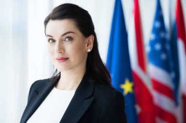 çekici büyükelçi bayrakların yanındaki kameraya bakıyor 