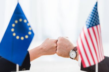 selective focus of diplomats bumping fists near flags of usa and european union clipart
