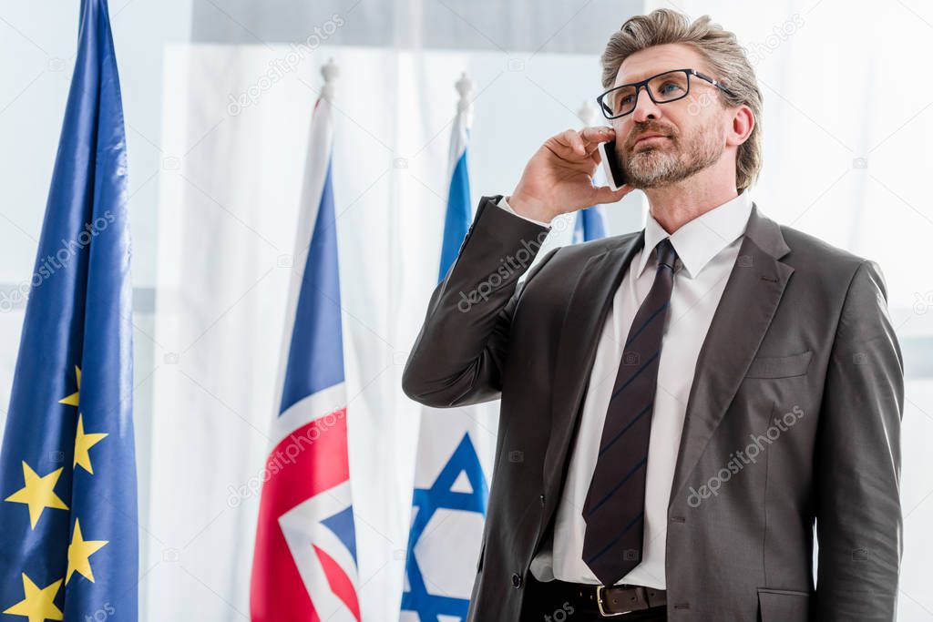 handsome bearded diplomat talking on smartphone near flags 