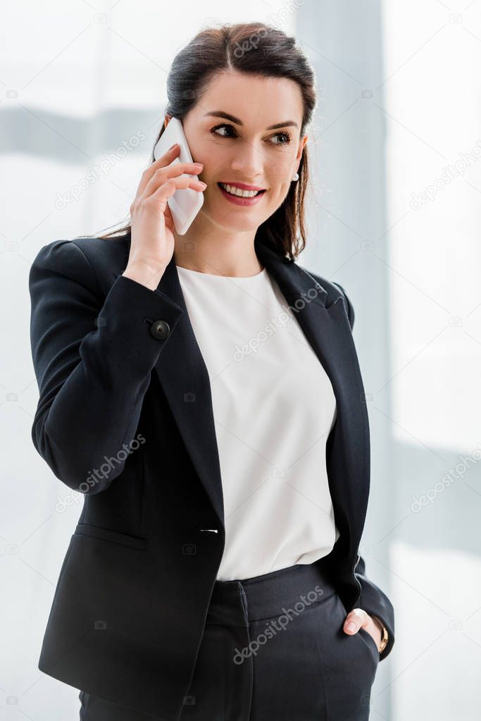 attractive diplomat standing with hand in pocket and smiling while talking on smartphone 