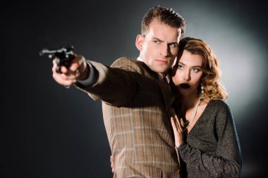 attractive woman standing near armed gangster with gun on black  clipart