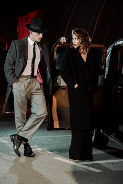handsome gangster standing with hand in pocket and looking at attractive woman near car  clipart