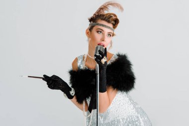 Retro mikrofona dokunurken şarkı söyleyen çekici bir kadın ve gri üzerinde izole edilmiş sigara tutacağı tutan bir kadın. 