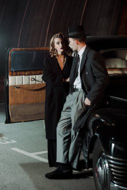 handsome gangster in hat looking at attractive woman near retro car  clipart