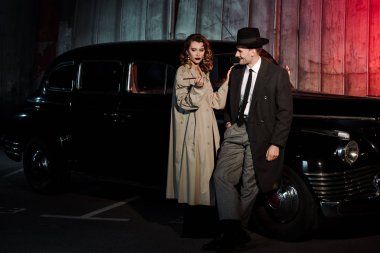 Şapkalı mutlu gangster, retro arabanın yanında sigara içen kadına bakıyor.