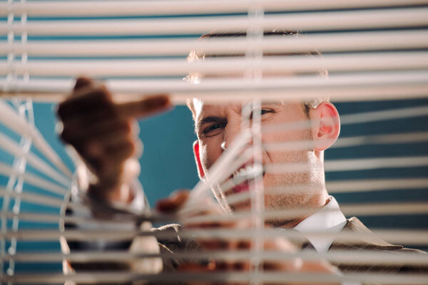 selective focus of emotional gangster showing middle finger through window blinds 