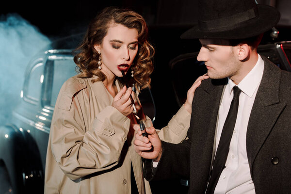 handsome gangster in hat holding lighter near attractive woman smoking near car on black with smoke 