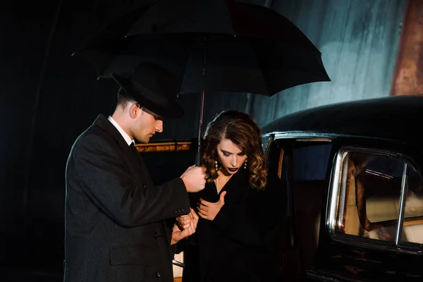 戴帽子的英俊匪徒拿着雨伞靠近女人和复古汽车 — 图库照片
