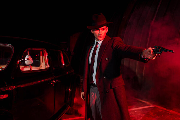  dangerous man in coat and hat holding gun near retro car 