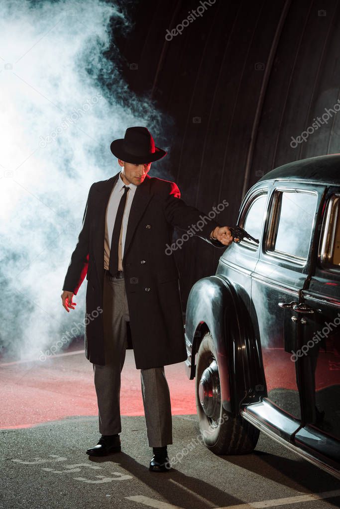 handsome gangster in hat holding gun near retro car and smoke