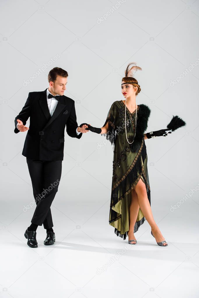 stylish man in suit and attractive woman holding hands and dancing on grey