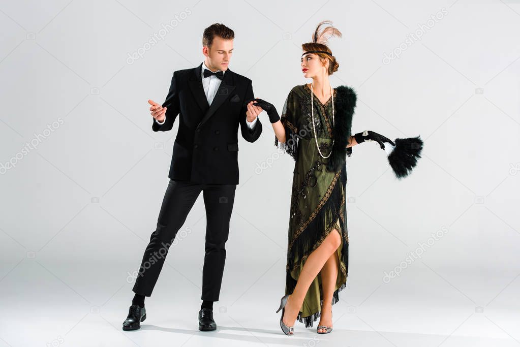 stylish man in suit and aristocratic woman holding hands while dancing on grey
