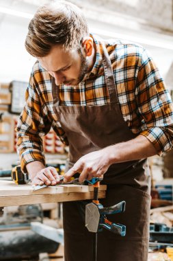 bearded carpenter in brown apron carving wood in workshop  clipart