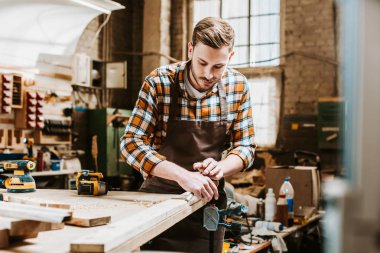 selective focus of bearded woodworker holding chisel while carving wood in workshop  clipart