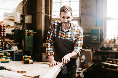 selective focus of happy bearded carpenter carving wood in workshop  clipart