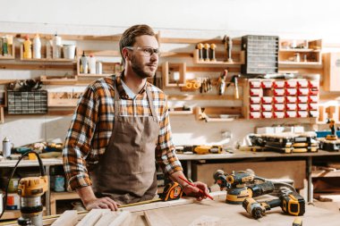 handsome carpenter in goggles standing near table and equipment  clipart