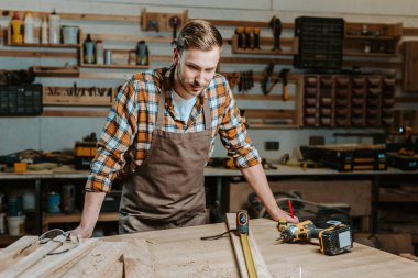 handsome woodworker looking at table with wooden planks and measuring tape  clipart
