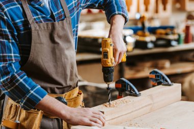 cropped view of carpenter in apron holding hammer drill near wooden planks clipart