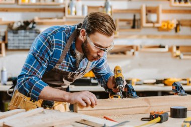 selective focus of carpenter in safety glasses and apron holding hammer drill and blowing on sawdust near wooden plank  clipart