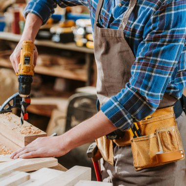 cropped view of carpenter in apron holding hammer drill near wooden plank  clipart