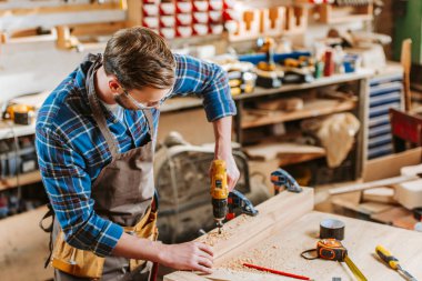 woodworker in goggles and apron holding hammer drill near wooden planks clipart