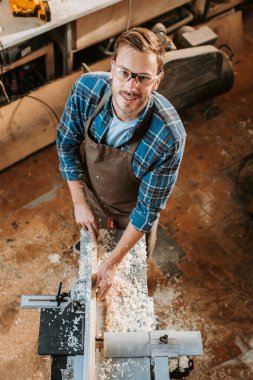 top view of happy carpenter holding plank near circular saw in carpentry shop  clipart