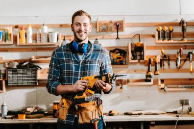 happy bearded carpenter holding hammer drill in workshop 