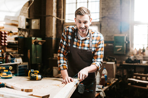 selective focus of happy bearded carpenter carving wood in workshop 