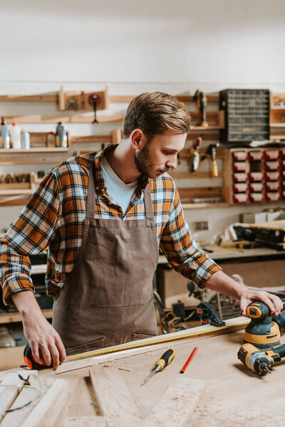 handsome carpenter in brown apron measuring wooden plank 