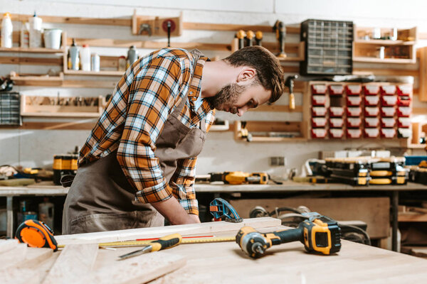 selective focus of bearded carpenter in brown apron measuring wooden plank 