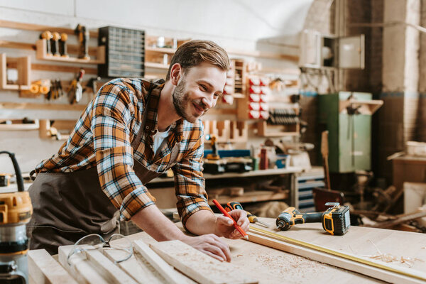 selective focus of happy woodworker holding pencil near wooden plank and measuring tape 