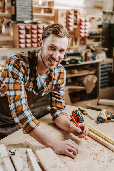 cheerful woodworker holding pencil near wooden plank and measuring tape 