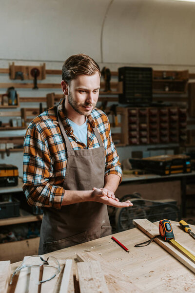 handsome woodworker standing near table with wooden planks and measuring tape 