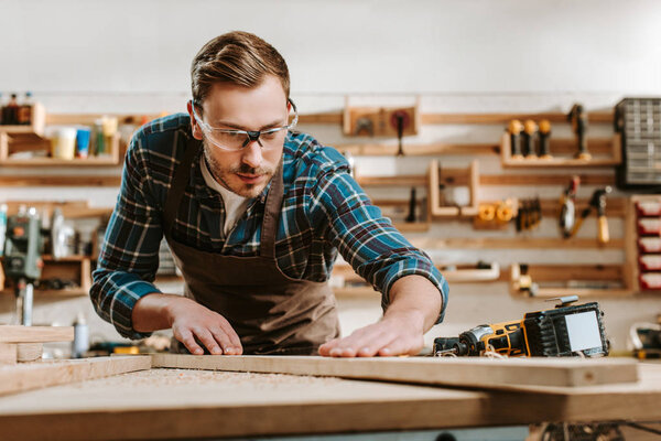 selective focus of handsome carpenter in goggles touching wooden plank 