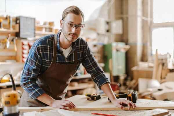 selective focus of handsome carpenter in goggles standing near wooden plank on table 