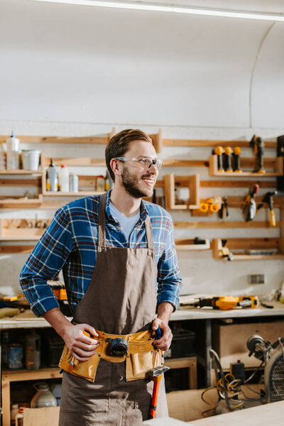 happy bearded carpenter in goggles touching tool belt 