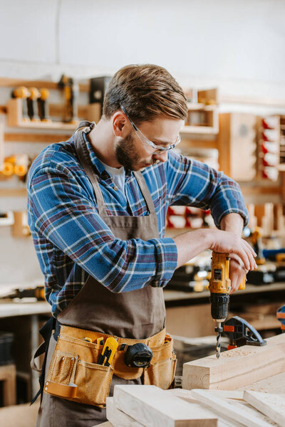 bearded carpenter in goggles and apron holding hammer drill near wooden planks