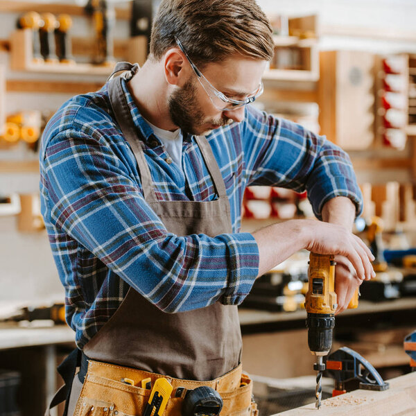 bearded carpenter in goggles and apron holding hammer drill near wooden plank