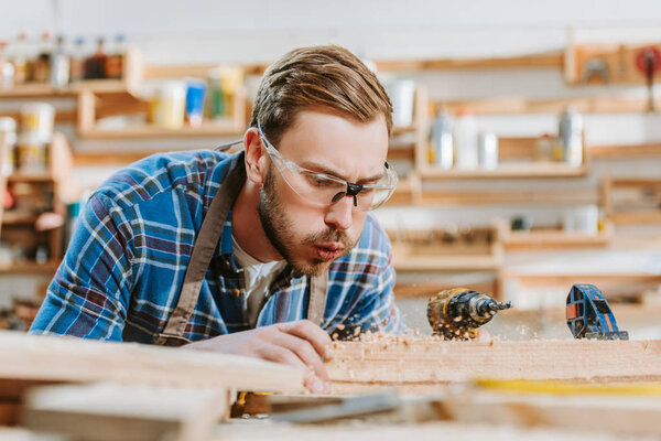 selective focus of carpenter in goggles holding hammer drill and blowing on sawdust near wooden plank 