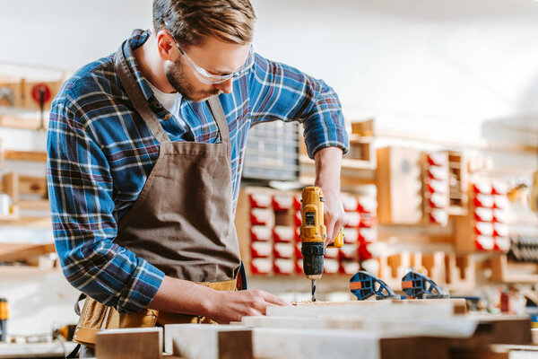 selective focus of woodworker in goggles and apron holding hammer drill near wooden planks
