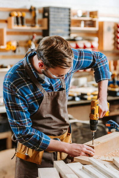 selective focus of woodworker in apron holding hammer drill near wooden planks