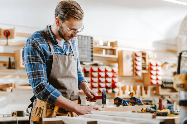 selective focus of carpenter in goggles holding hammer near wooden dowel in workshop 