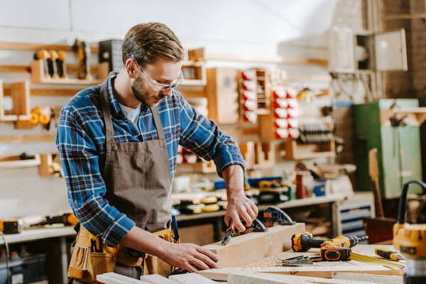 selective focus of bearded carpenter in goggles holding pliers near wooden dowel 