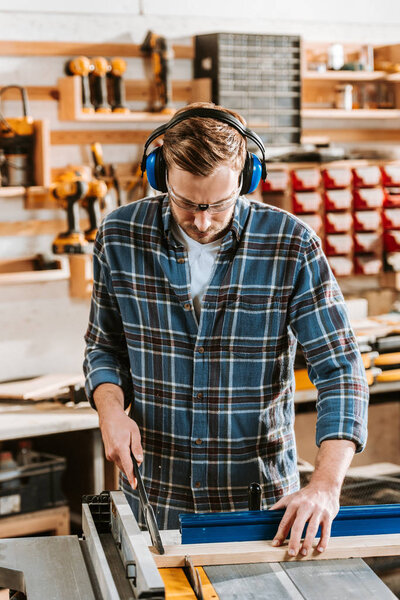 handsome carpenter in protective headphones holding plank near circular saw