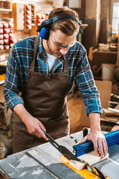 bearded woodworker in protective headphones and apron holding plank near circular saw