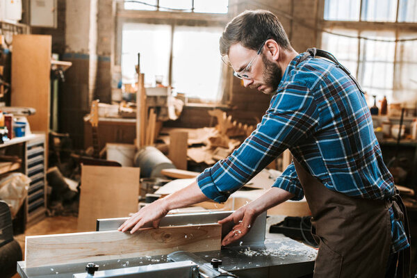 side view of bearded woodworker in apron holding plank near circular saw