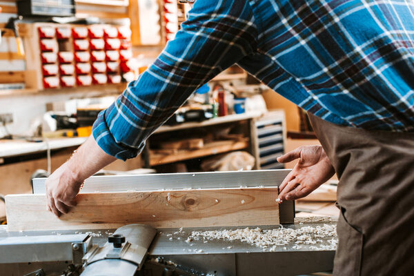 cropped view of woodworker holding plank near circular saw in carpentry shop 
